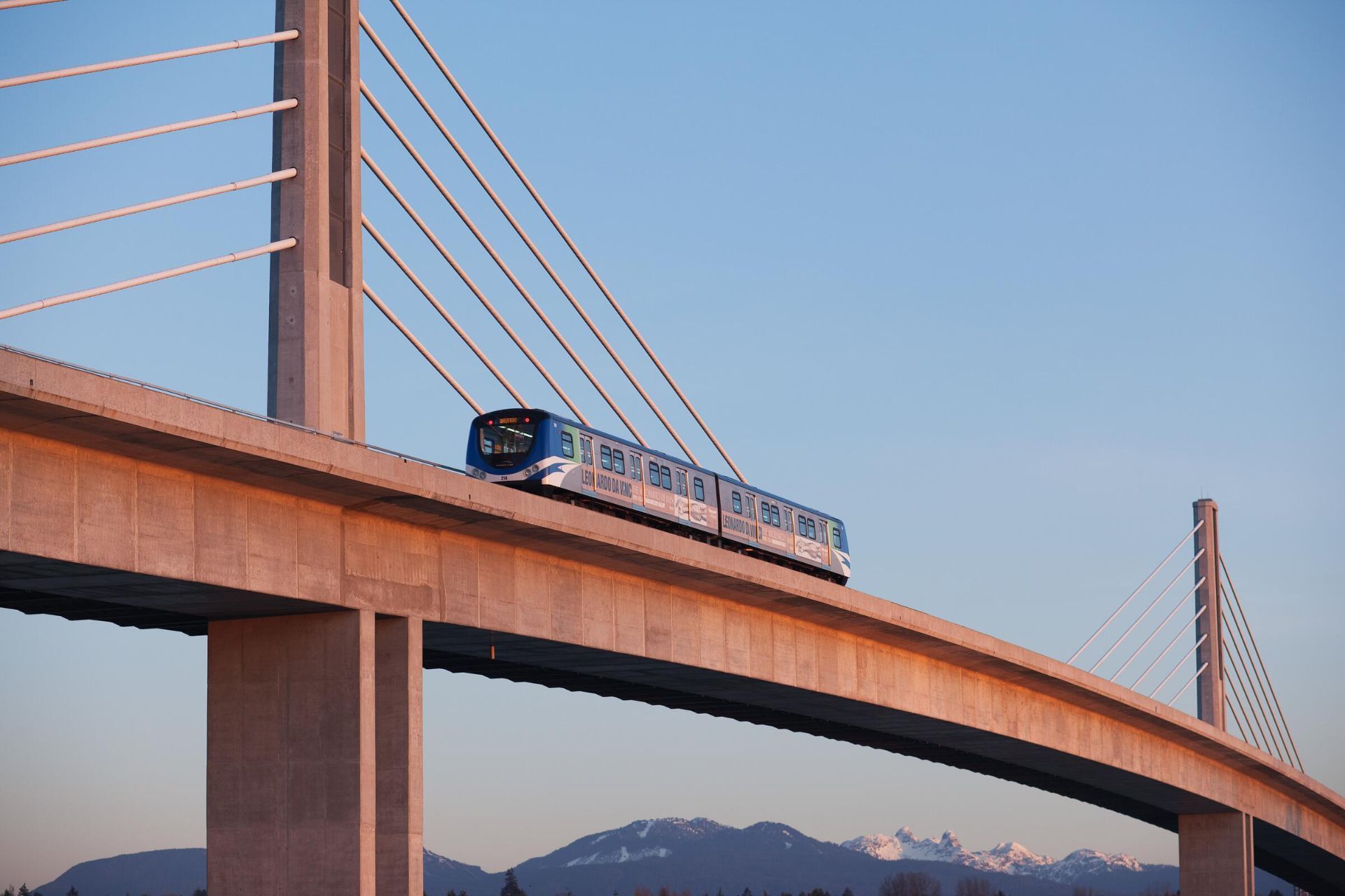 A Canada Line SkyTrain crossing a cable-stayed bridge at sunset, with the North Shore mountains behind